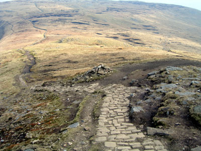 Path branches off the cobbles to the right, just before the cairn